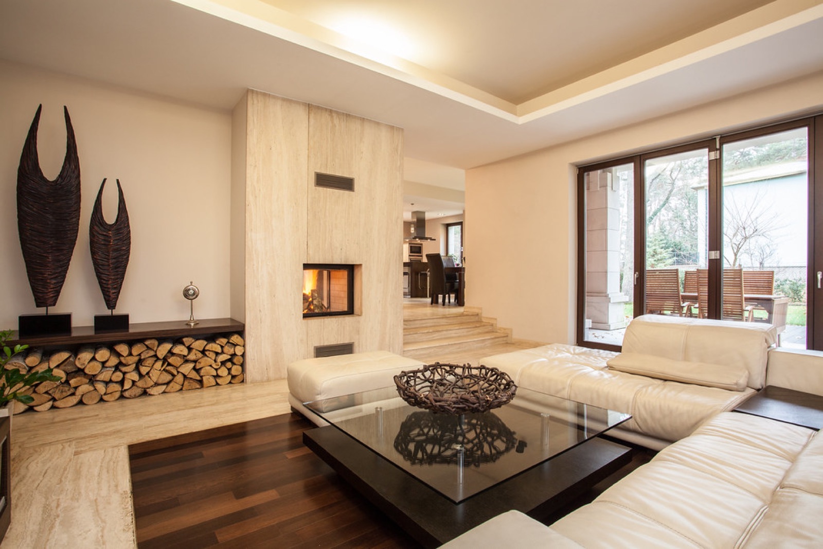 A bright modern living room with a cream sofa, a glass table, a travertine wall and doors open to a garden
