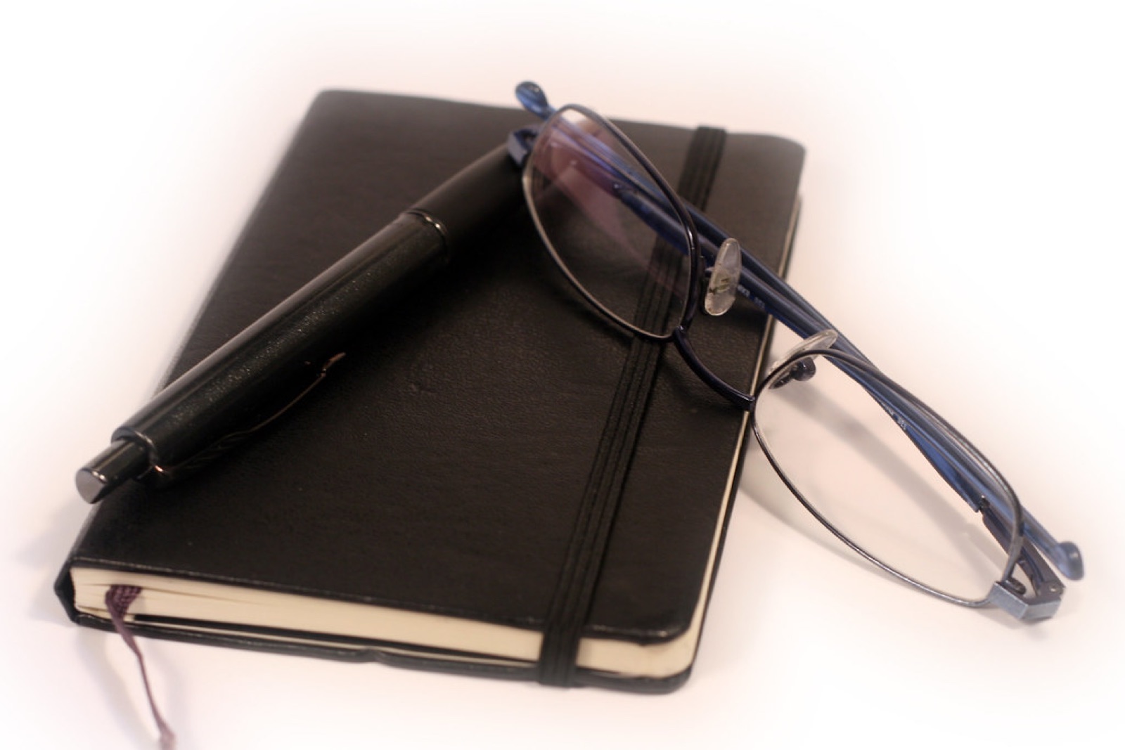 A closed black notebook with a pen and a pair of reading glasses resting on it